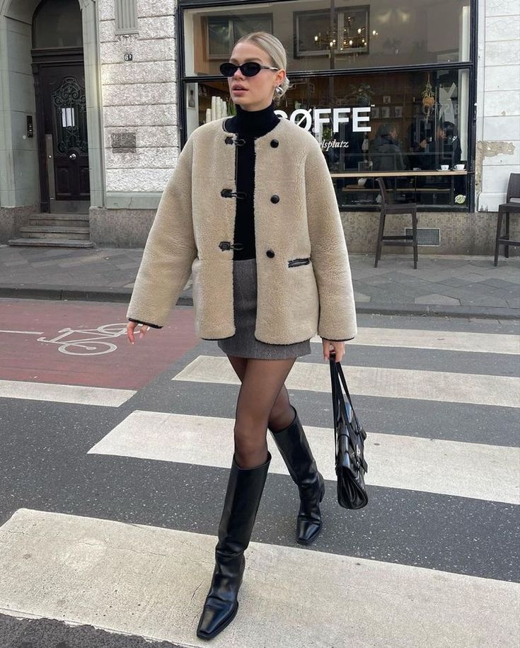 Linda usando óculos de sol, cabelo preso em coque, blusa de gola preta, casaco de lã, minissaia plissada, meia-calça de nylon preta e bota de cano médio de bico quadrado com bolsa preta