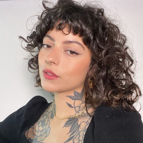 Mulher branca com blusa preta, cabelo cacheado escuro e maquiagem com delineado fino e longo