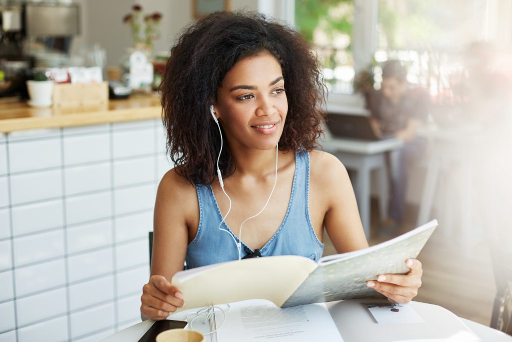 Mulher negra, jovem e sorridente, sentada, lendo um livro com fones de ouvido