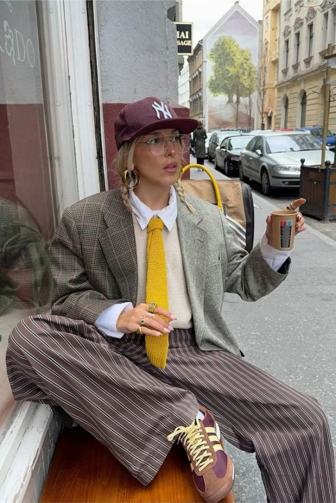 Sofia Coelho usando boné, gravata, camisa branca, pullover, blazer xadrez, calça de listra e tênis Adidas no estilo vovôcore