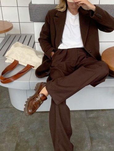Mulher sentada com pernas sobre a outra, usando boné em cor bege, camiseta branca, blazer marrom e calça de alfaiataria marrom.