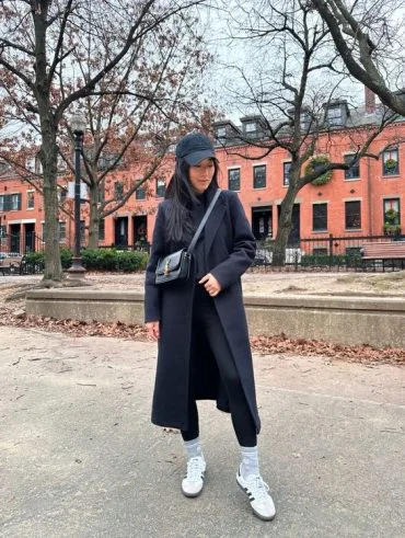 Mulher jovem e com cabelo escuro, usando boné preto, look all black, legging e Adidas Samba.