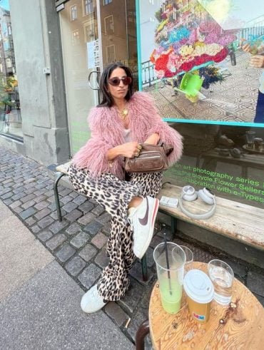 Mulher com cabelo escuro e curto, sentada em banco, usando óculos de sol, casaco rosa claro de pelugem, calça de oncinha e tênis da Nike.