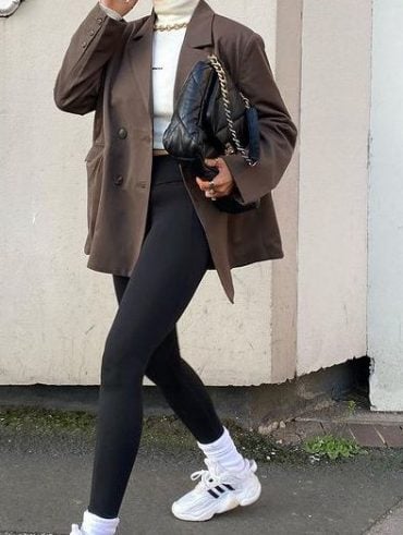 Mulher usando coque e óculos de sol, blusa de inverno com gola em tonalidade branca, blazer marrom, legging com meia alta e tênis.