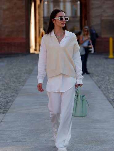 Mulher jovem, magra e de pele clara. Ela está vestindo um look todo branco com colete de tricot.