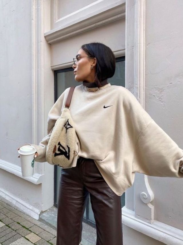 Mulher com cabelo moreno, usando blusa bege, bolsa no ombro e calça de couro em marrom., segurando copo de café.
