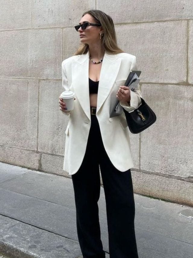 Mulher jovem, usando óculos de sol, blazer branco, calça de alfaiataria em preto, tênis branco e segurando uma bolsa pequena em preta.