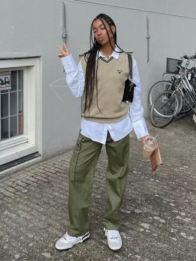Mulher negra, usando colete em tricô na cor bege, com bolsa no ombro em cor preta, calça cargo em verde militar e tênis branco.
