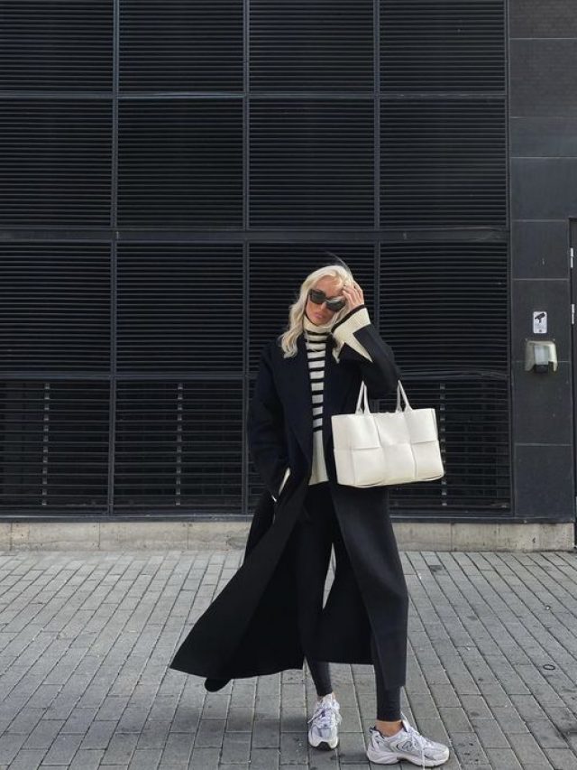 Mulher jovem e loira, usando óculos de sol, sobretudo preto, legging preta, tênis e segurando uma bolsa branca.