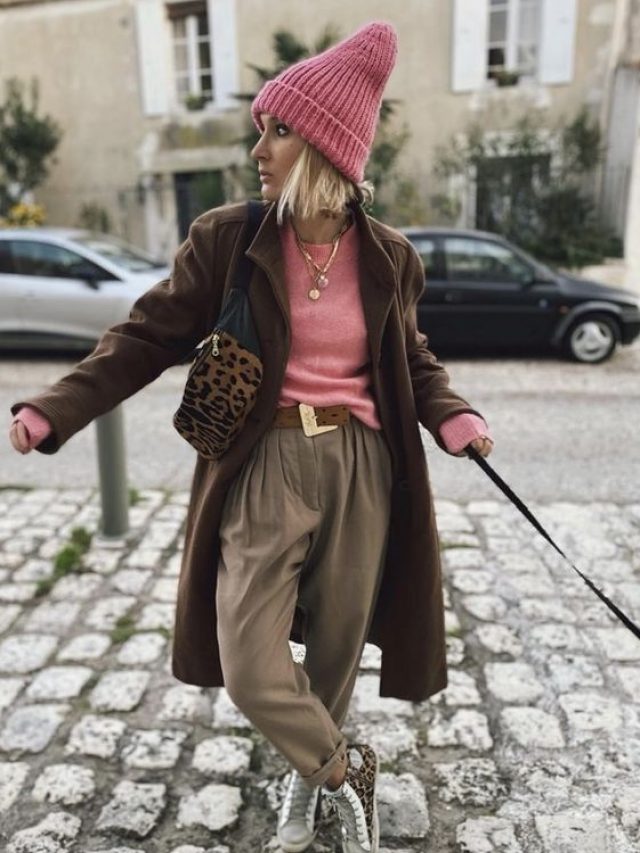 Mulher jovem e com cabelo loiro e curto, usando blusa rosa, touca rosa e calça de alfaiataria marrom claro com cinto.