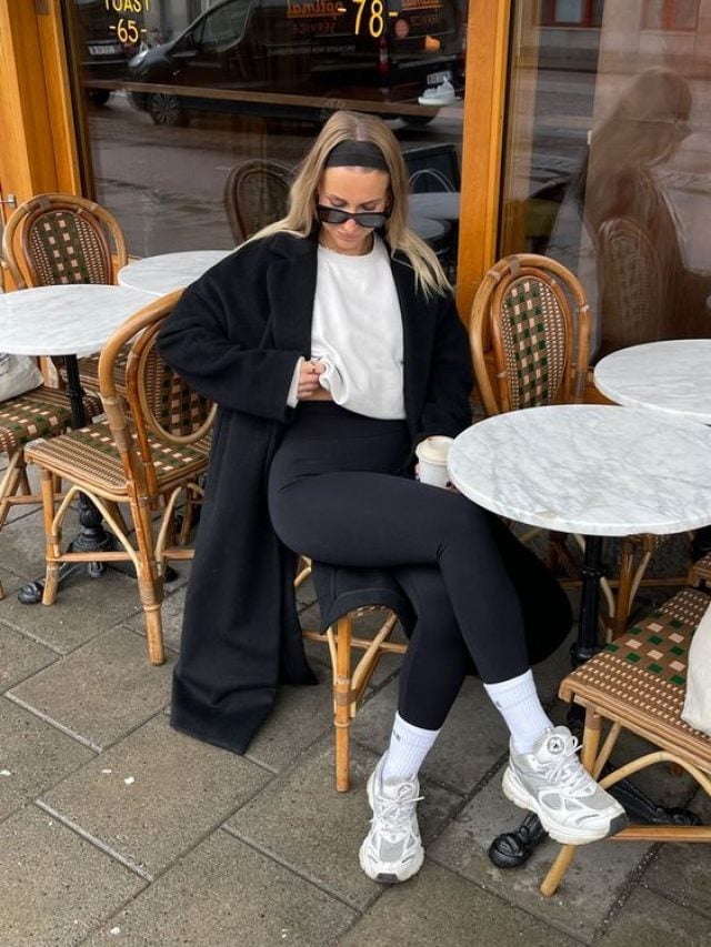 Mulher jovem e loira, usando faixa no cabelo, blusa branca, sobretudo preto, legging com meia alta e tênis, sentada em cadeira em frente à mesa.