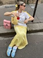 Mulher loira usando acessório de flor no cabelo, camiseta Adidas, saia midi acetinada amarelo manteiga, meia aparente, bolsa amarelo manteiga e tênis Adidas azul claro