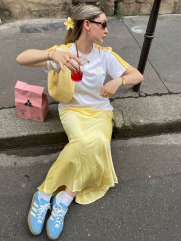Mulher loira usando acessório de flor no cabelo, camiseta Adidas, saia midi acetinada amarelo manteiga, meia aparente, bolsa amarelo manteiga e tênis Adidas azul claro
