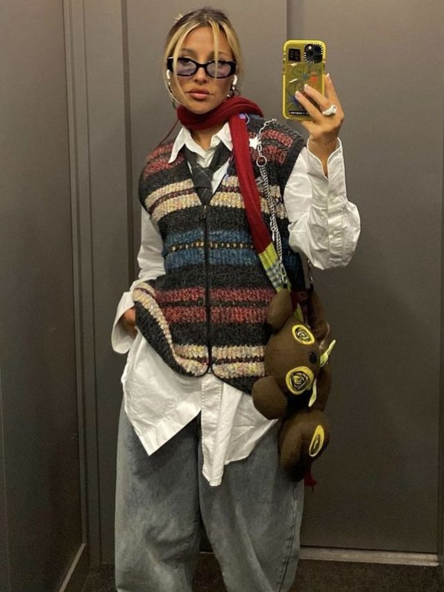 Mulher jovem branca usando óculos de sol, camisa branca, gravata, pullover, calça jeans oversized e mochila divertida no estilo vovôcore