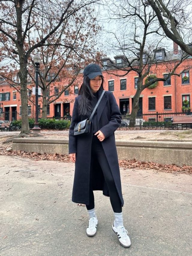 Mulher jovem e com cabelo escuro, usando boné preto, look all black, legging e Adidas Samba.