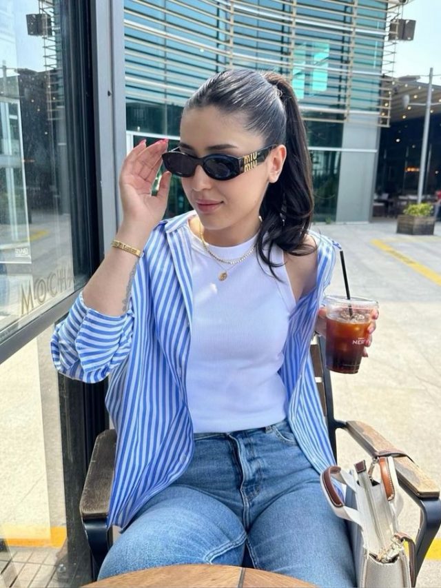 Mulher jovem de pele clara. Ela está sentada e veste camisa listrada azul, regata branca e short jeans.