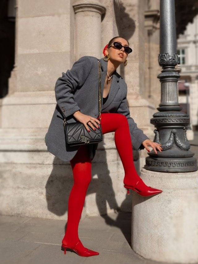 Mulher jovem, usando óculos de sol, com uma das pernas levanta, usando blazer cinza e meia-calça vermelha, com uma bolsa de tonalidade preta em um dos ombros.