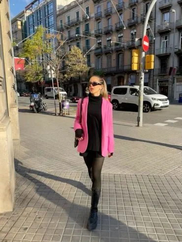 Mulher loira com cabelo preso, usando óculos de sol, blusa preta, blazer rosa, short preto, meia-calça e bota.