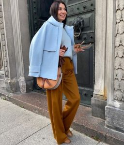 Mulher jovem e com cabelo moreno, usando casaco de lã batida em azul claro e calça em cor amarelo queimado.