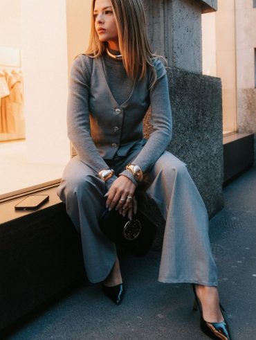 Mulher jovem e loira, sentada e usando look monocromático com calça de alfaiataria cinza e braceletes dourados.