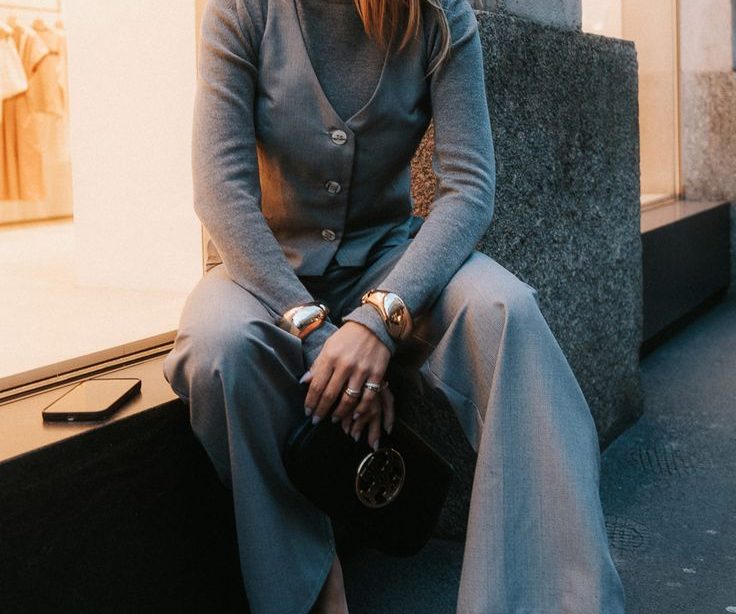 Mulher jovem e loira, sentada e usando look monocromático com calça de alfaiataria cinza e braceletes dourados.