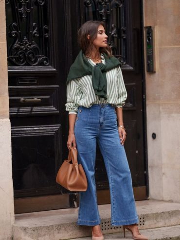 Mulher jovem de pele clara. Ela está parada na frente de uma porta e veste roupas casuais.