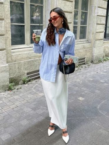 Mulher jovem usando look elegante com camisa de manga longa e saia de cetim. Ela está segurando um copo de plástico com bebida verde.
