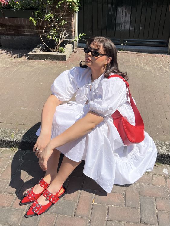 Mulher jovem e de pele clara. Ela está sentada usando vestido branco, bolsa vermelha e sapatilha cm tachas. 