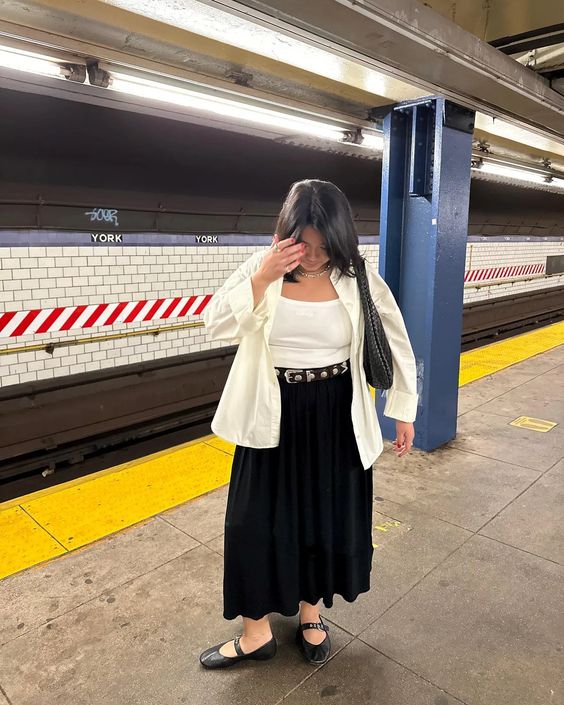 Mulher jovem. Ela usa blusa e casaco claro, saia longa preta e sapatilha. 