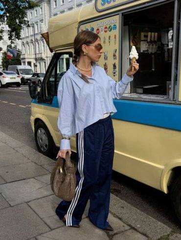 Mulher jovem com cabelo preso em coque, usando óculos de sol, segurando uma casquinha com sorvete, usando camisa azul e calça Adidas azul marinho, segurando uma bolsa.