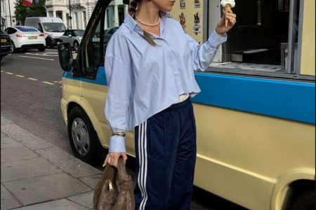 Mulher jovem com cabelo preso em coque, usando óculos de sol, segurando uma casquinha com sorvete, usando camisa azul e calça Adidas azul marinho, segurando uma bolsa.