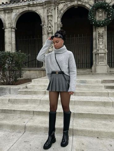 Mulher jovem e negra, usando tricô cinza, saia de pregas cinza, meia-calça e botas de canos altos.