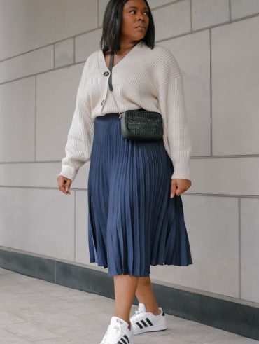 Mulher midisize e negra, com cabelo curto, usando casaco de tricô branco, saia plissada midi em azul e tênis.
