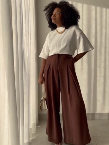 Mulher negra, com cabelo black power, usando camiseta de gola branca e calça pantalona em cor marrom.