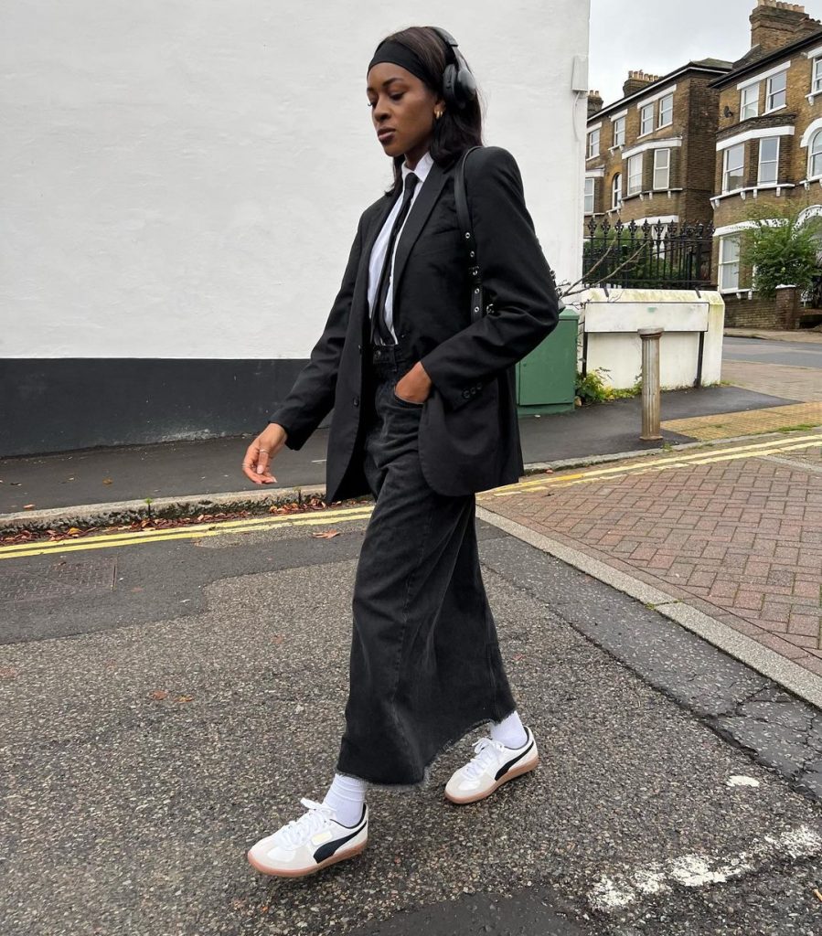 Georgia usando faixa de cabelo, headphone, blazer preto, camisa branca, gravata, saia longa jeans, meia branca e tênis Puma Palermo branco com preto