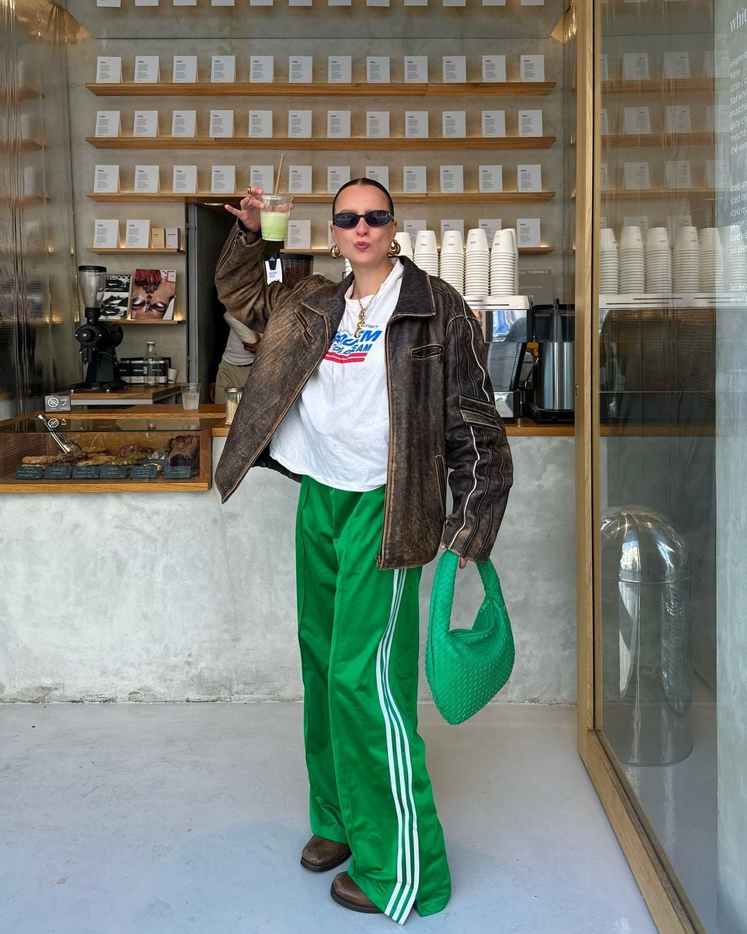 Mulher jovem e magra com pele clara. Ela veste jaqueta estonada, camiseta branca com estampa e calça adidas verde.