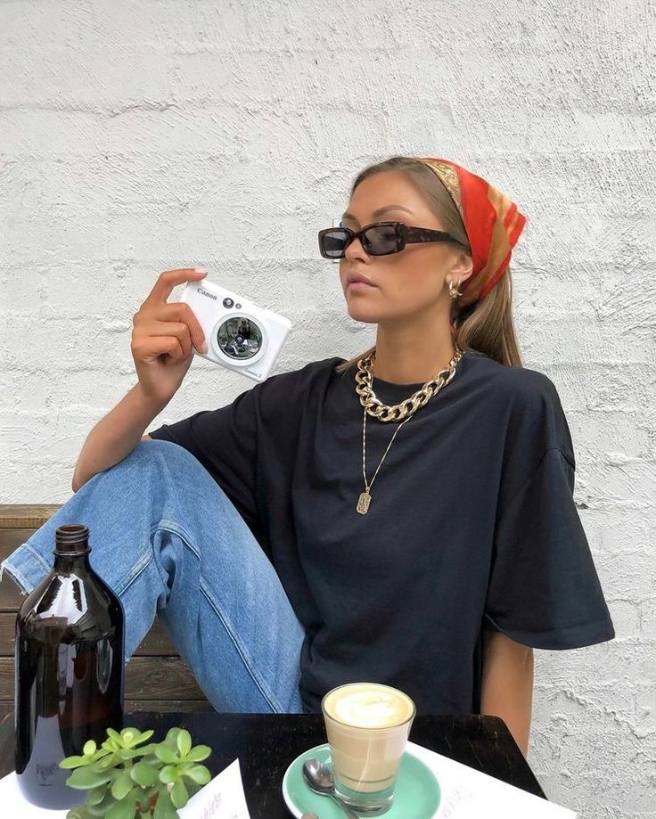 Mulher jovem branca com lenço no cabelo liso. Ela usa óculos de sol, acessórios dourados, camiseta preta básica e calça jeans