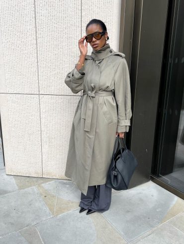 Marilyn com trench coat, óculos de sol de lente transparente colorida, calça de alfaiataria cinza, bolsa preta e sapato de bico fino