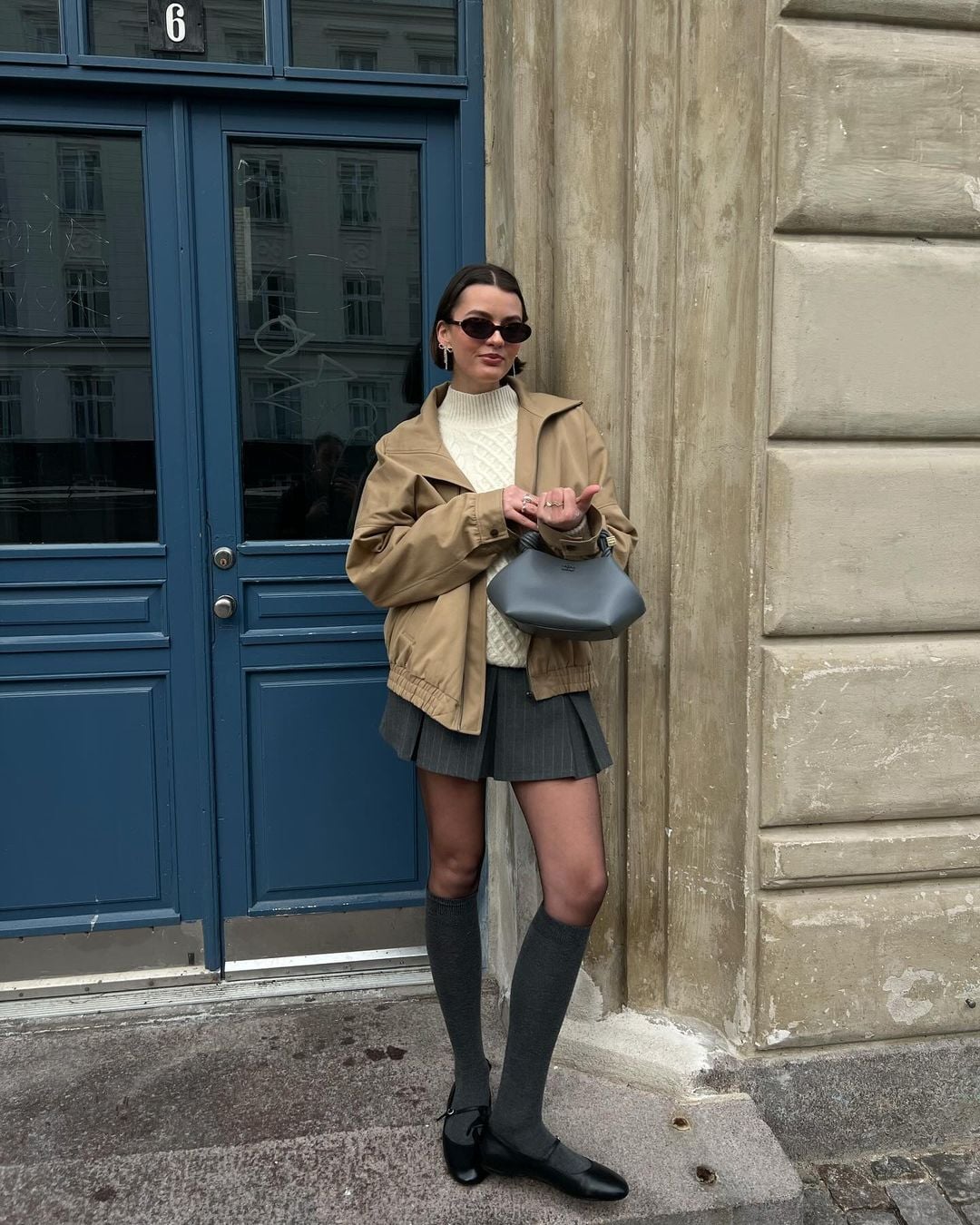 Mary usando óculos de sol minimalista, blusa de gola alta off-white, jaqueta, minissaia de pregas cinza, meia três quartos cinza e sapatilha de Mary Jane