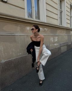 Mary usando óculos minimalista, blusa preta sem alça, calça risca de giz off-white e sapatilha de bailarina