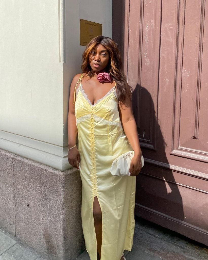 Nnenna usando slip dress amarelo-manteiga, acessório de flor no pescoço e bolsa branca