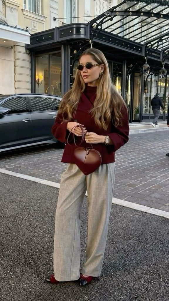 Mulher jovem branca usando óculos de sol, casaco vermelho cereja, bolsa de coração vermelho cereja, pantalona de alfaiataria e sapatilha bicolor preto com vermelho cereja