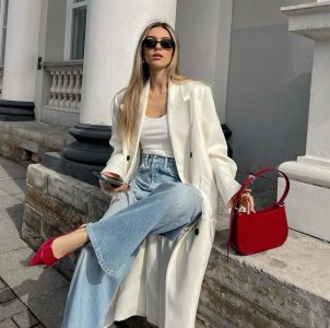 Mulher jovem loira usando sobretudo off-white, calça jeans e slingback vermelho intenso