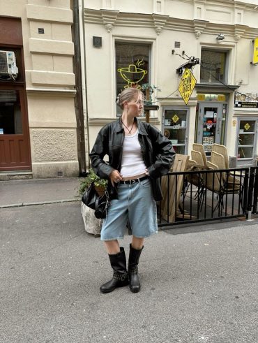 Mulher jovem de pele clara e magra. Ela usa uma regata branca, jaqueta preta, bermuda jeans e biker boots.