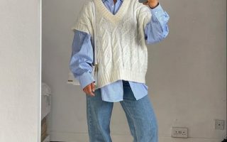 Mulher jovem e loira, com cabelo preso e trança, usando óculos de sol, camisa oversized em cor azul, colete branco de alfaiataria em sobreposição e calça jeans.