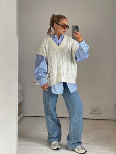 Mulher jovem e loira, com cabelo preso e trança, usando óculos de sol, camisa oversized em cor azul, colete branco de alfaiataria em sobreposição e calça jeans.