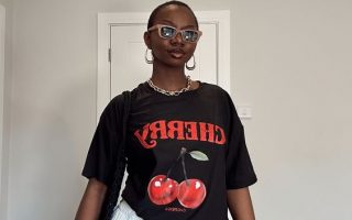 Mulher jovem e magra com pele escura. Ela usa camiseta preta estampada e saia branca.