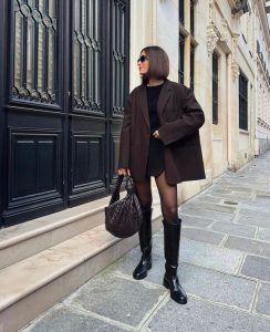 Mulher jovem de cabelo curto, usando óculos de sol, blazer marrom, meia-calça preta e bota de cano alto.