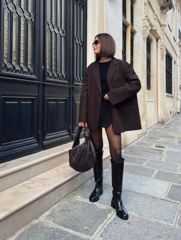 Mulher jovem de cabelo curto, usando óculos de sol, blazer marrom, meia-calça preta e bota de cano alto.