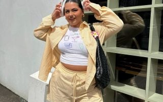 Mulher jovem de pele clara e cabelo escuto. Ela usa conjunto amarelo manteiga de camisa e bermuda, e blusa branca estampada.